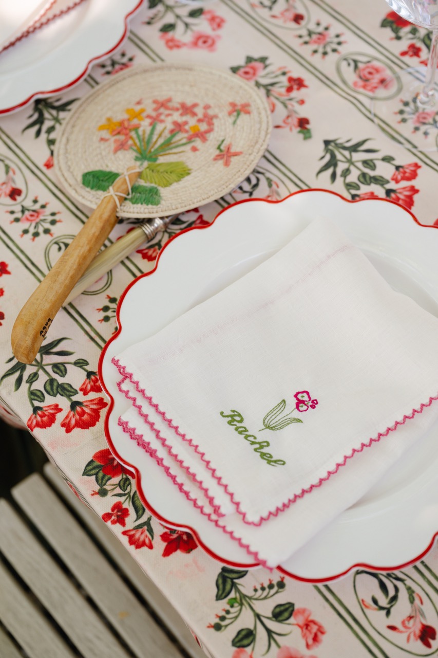 Close-up of an elegant place setting at the Agua by Agua Bendita x CAKE brand activation, featuring a floral embroidered napkin, scalloped dinner plate, hand-painted fan, and botanical table linens for an outdoor Hamptons dinner.